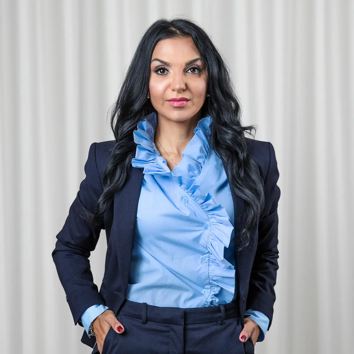 Kvinna med långt mörkt hår i blå kostymjacka och ljusblå blus står mot en neutral bakgrund